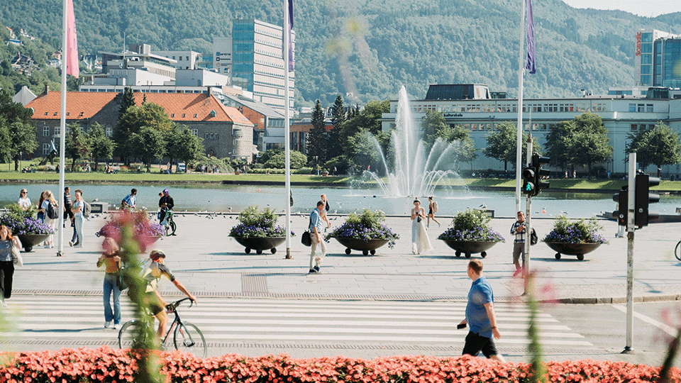 Pilt "Festplassen" Bergeni keskuses. Pildil on inimesed, kes kõnnivad ja sõidavad jalgrattaga üle väljaku ja ülekäigurada. Plats on sillutatud kiviga, taustal on vesi. Vees on vees purskkaev, mis tekitab pildil liikumist.